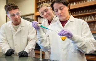 University of Mary SURVE students Ethan Emineth (left), Grace Dahl (middle), and Mariapocs Ruiz Martinez (right). Mariapocs “Maria” is demonstrating how media is removed from a culture flask so that the number of cells in the culture can be measured. She is using aseptic technique in the presence of a Bunsen burner so that the culture media does not become contaminated. Credit: Mike McCleary/University of Mary